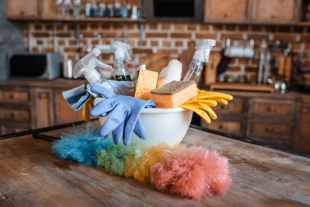 Assorted cleaning supplies in a bowl on a wooden table in a rustic kitchen, ready for move-out cleaning.”
